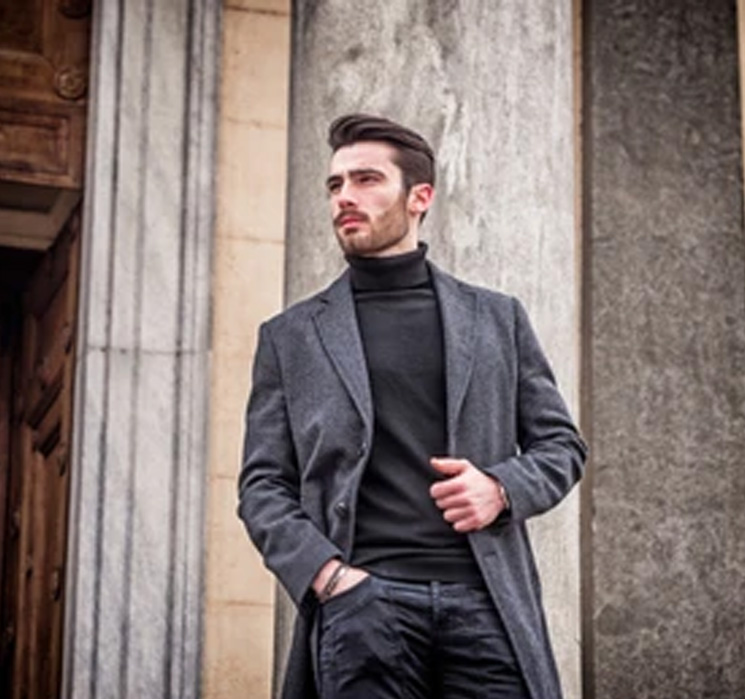 Man in a gray coat and black turtleneck standing outdoors.